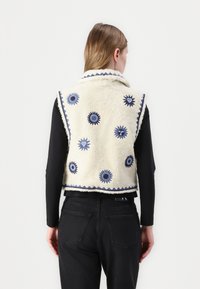 Fleeceväst i krämfärg med blå broderade blommönster, krage och kontrasterande detaljer. Bakvy, ihop med svarta jeans.