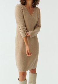 Robe-pull en tricot beige avec encolure en V, manches longues, poignets et ourlet côtelés. Texture douce, silhouette ajustée et longueur aux genoux.