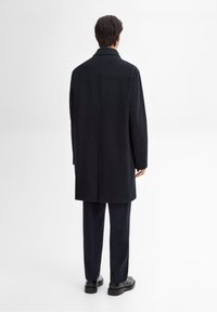 Homme debout de dos, portant un long manteau noir, un pantalon noir et des chaussures noires sur un fond blanc uni.
