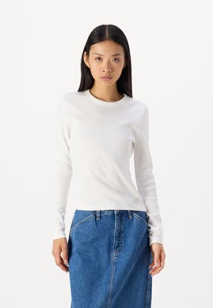 Maglietta bianca a maniche lunghe in rib liscio con scollatura rotonda, abbinata a una gonna di jeans. La maglietta presenta una texture sottile e un piccolo dettaglio con etichetta.