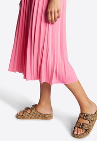 Personne portant une jupe plissée rose et des sandales à double boucle marron au design à motifs, marchant sur un fond blanc.