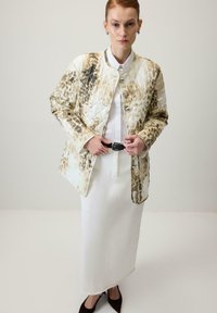 Nő, aki egy fekete övet igazít, bézs kígyómintás steppelt dzsekit, fehér inget, hosszú fehér szoknyát és fekete hegyes orrú cipőt visel egy sima hátterű képen.