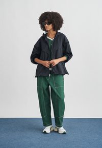 Μαύρο μπουφάν νάιλον με άνετη εφαρμογή και κουκούλα, φορεμένο πάνω από πράσινο τοπ. Πράσινο παντελόνι με λευκό ραφές, συνδυασμένο με λευκά αθλητικά παπούτσια.