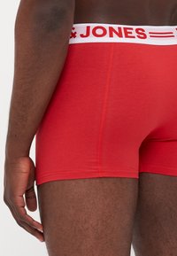 Rote Boxershorts aus einem glatten Material, mit einem weißen Taillenbund, auf dem der Text "JONES" in Rot steht. Nahaufnahme von der Seitenansicht.