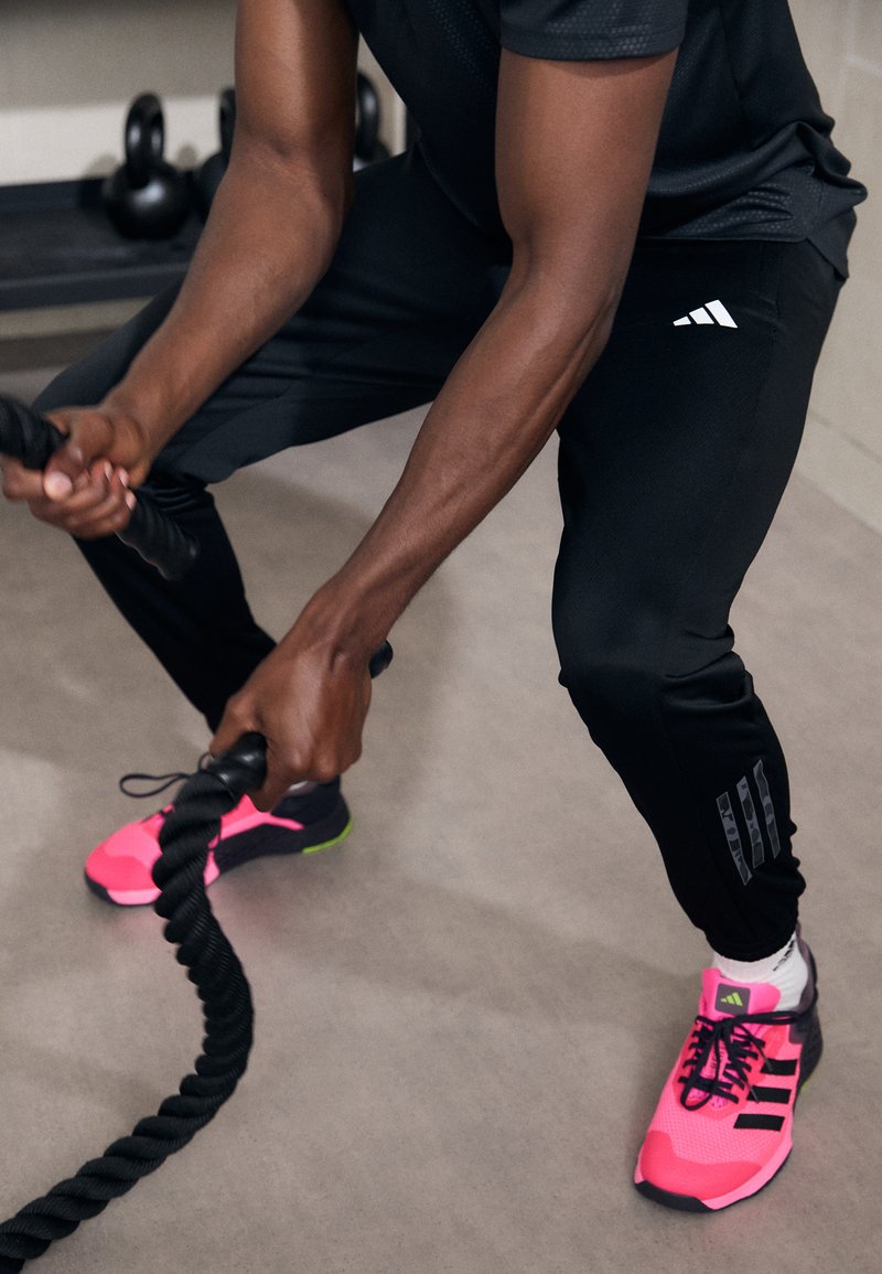 Pantalones deportivos negros con tres rayas blancas, asas de agarre negras y zapatillas deportivas de color rosa brillante con detalles en negro y suelas verdes.
