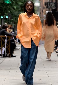 Modellen har på seg en oversized, knappened stråfarget skjorte og løse, mørkeblå denimjeans mens hun går på en bygate.