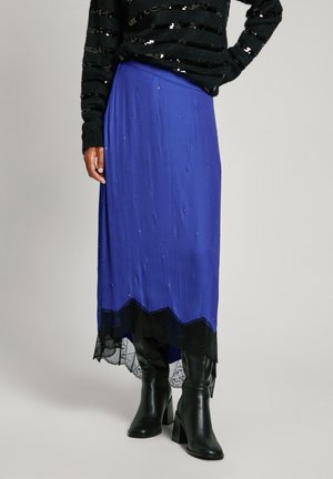 Jupe midi bleue avec des accents en perles et un ourlet en dentelle noire à volants, associée à des bottes noires à talons. Pull noir texturé porté par-dessus.