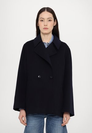 Manteau en laine marine avec un design à double boutonnage, grand col et manches larges. Présente des boutons noirs et une coupe droite. Porté sur une chemise légère.