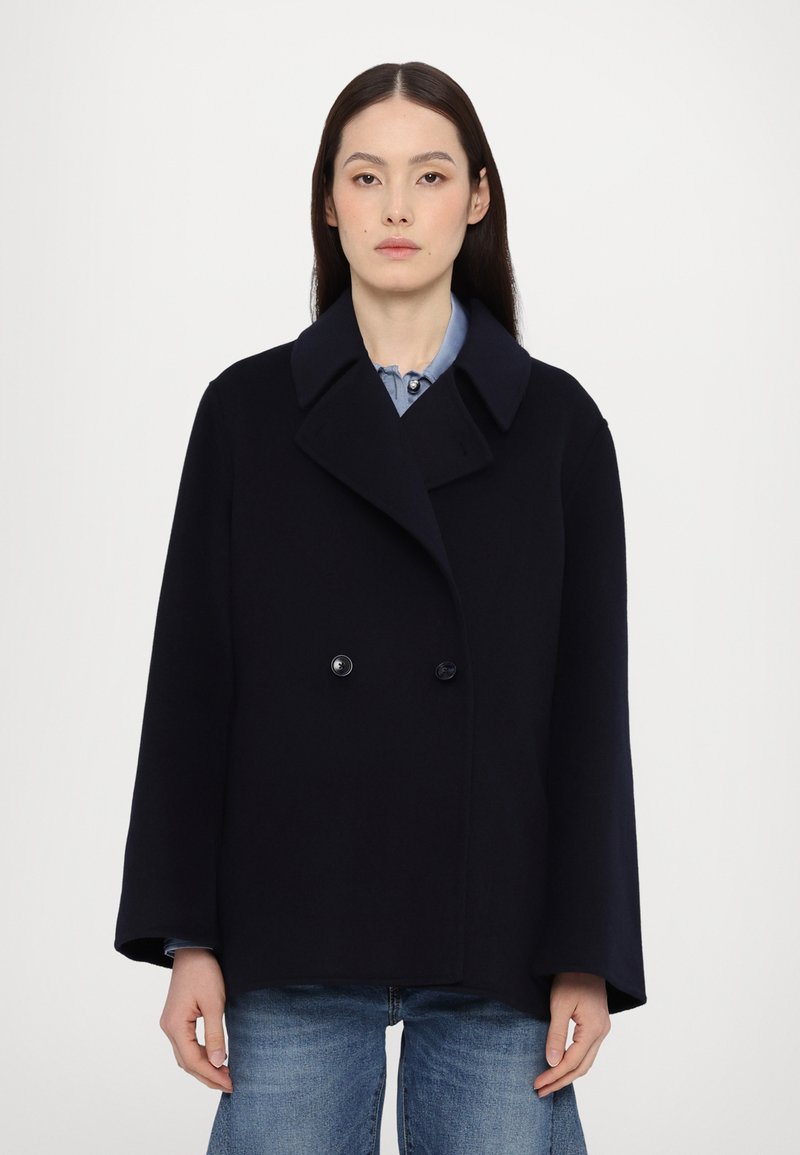Cappotto di lana blu navy con un design doppiopetto, colletto ampio e maniche larghe. Presenta bottoni neri e una linea dritta. Indossato sopra una camicia chiara.