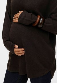 Mørkebrun strikket sweater med ribbede manchetter. Et træarmbånd pryder det ene håndled. Hånden hviler på en synlig gravid mave, der viser blødhed.