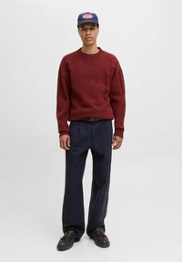 Pull en maille texturée bordeaux avec des poignets côtelés, associé à un pantalon navy ample et des chaussures marron foncé. La casquette arbore un patch logo.