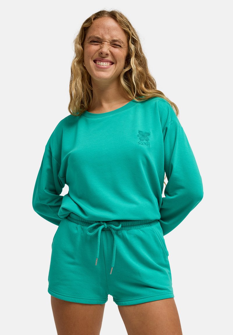 Türkisfarbenes Langarmshirt kombiniert mit passenden Shorts mit Kordelzug. Weicher Stoff, lockere Passform, mit gesticktem Logo auf der Brust.