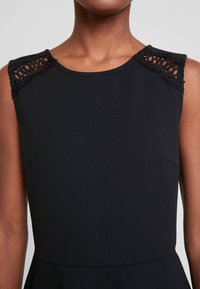 Czarna sukienka z gładkim, dopasowanym gorsetem oraz misternymi koronkowymi detalami na ramionach i rozkloszowanym dołem. Miękka faktura materiału.