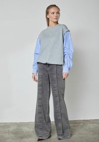 Szürke rövid ujjú pulóver, kék csíkos inggel rétegezve, széles szárú szürke farmernadrággal, melynek oldalán gombos részletek találhatók.