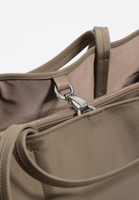 Beżowa torba typu tote wykonana z gładkiej tkaniny, wyposażona w srebrny zamek błyskawiczny i okrągłe elementy metalowe, z długimi, zaokrąglonymi uchwytami i minimalistycznym wzornictwem.