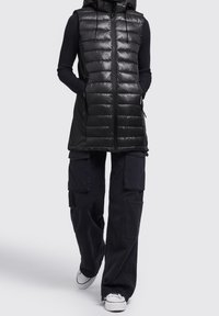 Czarna pikowana kamizelka z kapturem, z przeszywanym wzorem i przednim suwakiem. Nosi się ją na czarną bluzkę z długim rękawem i łączy z luźnymi spodniami typu cargo.