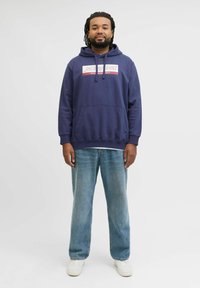 Mørkeblå hoodie med en frontlomme og hvidt logodetalje, parret med lyseblå jeans og hvide sneakers, stående mod en ensfarvet baggrund.