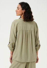 Bluză verde deschis cu mâneci lungi, detaliu cu pliu pe spate și mâneci rulabile. Fabricată dintr-un material texturat, cu o croială lejeră.