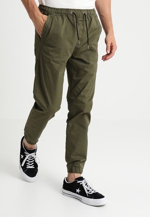 Hombre con pantalones jogger verde oliva con cordón y zapatillas negras con cordones blancos, de pie con una mano en el bolsillo.