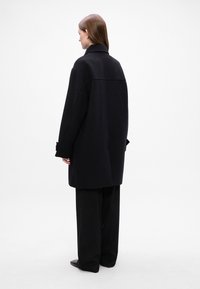 Cappotto nero oversize con colletto strutturato e maniche larghe. Tessuto liscio, dettagli di cucitura minimi. Abbinato a pantaloni neri larghi.