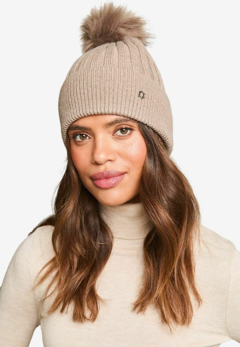 Beige knit beanie with ribbed texture, topped with a fluffy pom-pom. Features a small logo on the side.