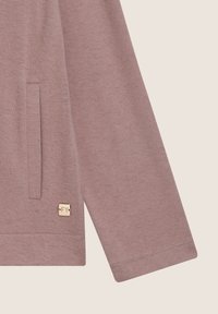 Maglietta a maniche lunghe di colore rosa con tasca laterale, orlo a coste e una piccola etichetta di marca sul lato, dal texture liscia.