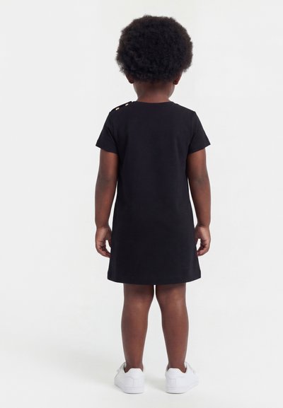 Enfant aux cheveux courts et bouclés, portant une robe noire à manches courtes et des baskets blanches, debout de dos sur un fond uni.