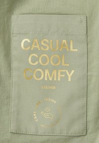 Zielony materiał z naszywaną kieszenią, na której znajduje się złoty napis: "CASUAL COOL COMFY", nazwa marki "S.OLIVER" oraz ikona instrukcji pielęgnacji.