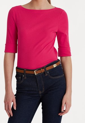 Person iført en knaldrosa trekvartærmet top med mørkeblå jeans og et brunt bælte mod en ensartet hvid baggrund.