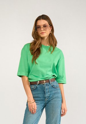Donna con i capelli lunghi e castani che indossa una maglietta verde oversize, jeans blu a vita alta, cintura marrone, occhiali da sole e gioielli d'oro, in piedi davanti a uno sfondo neutro.