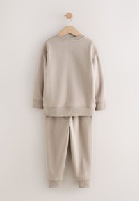 Felpa beige a maniche lunghe e pantaloni della tuta coordinati appesi su una gruccia di legno contro una parete chiara.