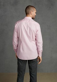 Helles rosa Hemd mit langen Ärmeln, Knopfleiste und abgerundetem Saum, kombiniert mit dunkelgrauen Hosen. Flache Textur, minimalistische Designdetails.