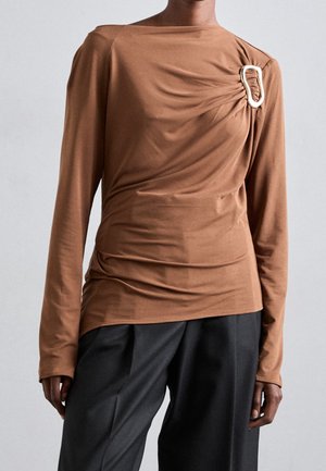 Brązowa bluzka z długim rękawem, materiał zebrany na prawym ramieniu, zabezpieczony dużą srebrną ozdobną klamrą, noszona z luźnymi czarnymi spodniami.