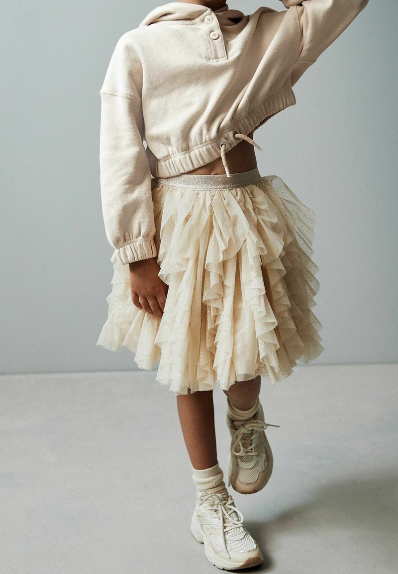 Enfant portant un sweat-shirt court beige clair, une jupe en tulle superposée, des chaussettes et des baskets épaisses, debout avec une jambe légèrement levée.