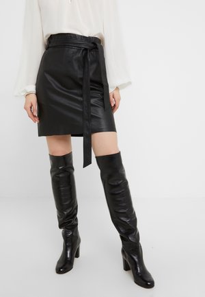 Femme portant une jupe portefeuille en cuir noir avec une ceinture nouée et des bottes à talons hautes en cuir noir jusqu'aux genoux, assorties à un chemisier blanc à manches longues.