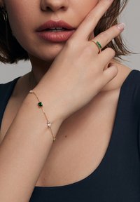 Złota bransoletka z zielonymi i przezroczystymi kamieniami szlachetnymi, z delikatnym łańcuszkiem. Dłoń modelki opiera się na twarzy, podkreślając biżuterię.