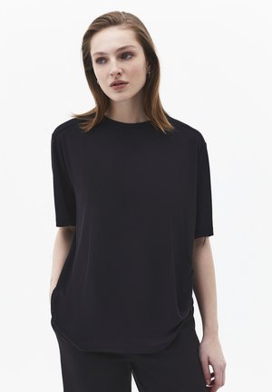 Ung kvinde med lysebrunt hår iført en løs sort kortærmet skjorte og sorte bukser foran en ensartet hvid baggrund.