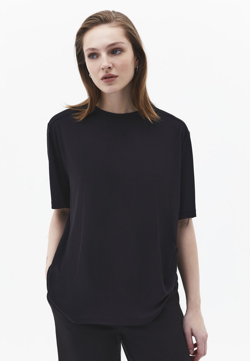 Mujer joven de cabello castaño claro, vistiendo una camisa negra de manga corta y suelta y pantalones negros, frente a un fondo blanco liso.