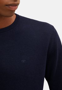 Granatowy sweter z okrągłym dekoltem wykonany z miękkiej, dzianinowej tkaniny. Dyskretne logo na piersi. Żebrowany dekolt i mankiety, gładka tekstura, dopasowany krój.