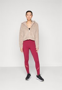 Veste à zip en polaire beige, associée à des leggings bordeaux taille haute avec des panneaux en mesh ; le modèle porte des chaussures de sport noires.