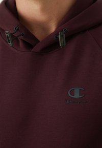 Hoodie bordeaux en tissu doux avec un grand cordon de serrage ajustable et un logo brodé noir sur la poitrine. Présente une texture lisse.