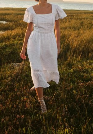 Femme marchant dans une haute herbe près de l'eau au coucher du soleil, portant une robe midi blanche en broderie anglaise et des sandales à lacets.