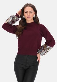 Bordeaux trui met geribbelde halslijn en lange mouwen, voorzien van pailletten en een metallic patroon bij de mouwen. Draag het met strakke zwarte broek.