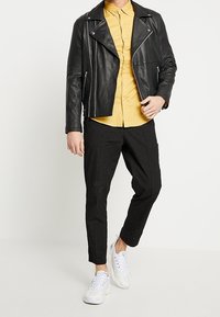 Mężczyzna w czarnej skórzanej kurtce na musztardowej koszuli, w czarnych dopasowanych spodniach i białych trampkach, stojący na tle białym.