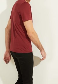 Hombre de pie de lado, vistiendo una camiseta corta ajustada de color burdeos y pantalones vaqueros negros contra un fondo claro y liso.