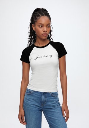 Femme avec les cheveux tressés portant un t-shirt blanc et noir à manches courtes avec le texte "juicy" et un jean bleu, debout devant un fond uni.