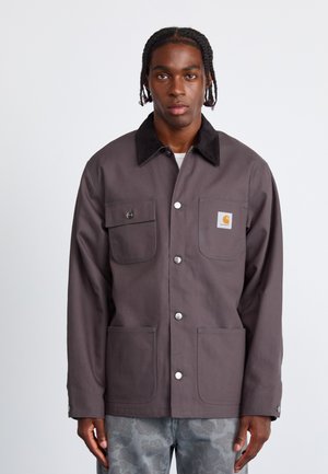 Gray canvas jacket with a black corduroy collar, two front pockets, and button closure. Features a small Carhartt logo on the chest pocket.