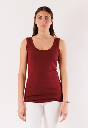 Donna con lunghi capelli castani, indossa una canotta color melograno e pantaloni bianchi, in piedi davanti a uno sfondo chiaro e uniforme.