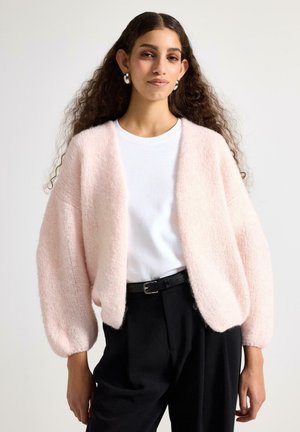 Kvinde med langt krøllet hår iført sølvfarvede hoops, en lyserød blød cardigan, hvid t-shirt, sorte bukser og sort bælte mod en ensfarvet baggrund.