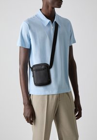Borsa a tracolla nera in pelle liscia con zip, indossata sopra una polo azzurra. Il modello indossa anche pantaloni beige.